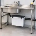 Filtre à plâtre installé sous un évier en inox, avec une tuyauterie d’évacuation Geberit.
